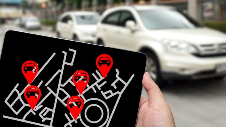Em primeiro plano, uma mão segurando um ipad e na tela mostrando uma ilustração de geolocalização de carros. No segundo plano, carros.