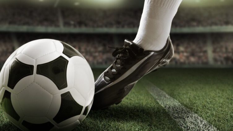 Imagem de uma pessoa usando chuteiras e chutando uma bola de futebol