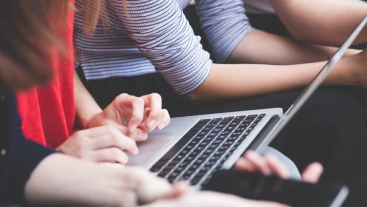 Imagem de diversas pessoas participando de uma reunião. Uma delas está mexendo em um laptop, enquanto outros mexem no celular