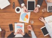 Imagem de uma reunião em ambiente corporativo com as pessoas utilizando laptops, tablets, celulares e gráficos impressos em papel