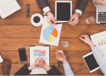 Imagem de uma reunião em ambiente corporativo com as pessoas utilizando laptops, tablets, celulares e gráficos impressos em papel