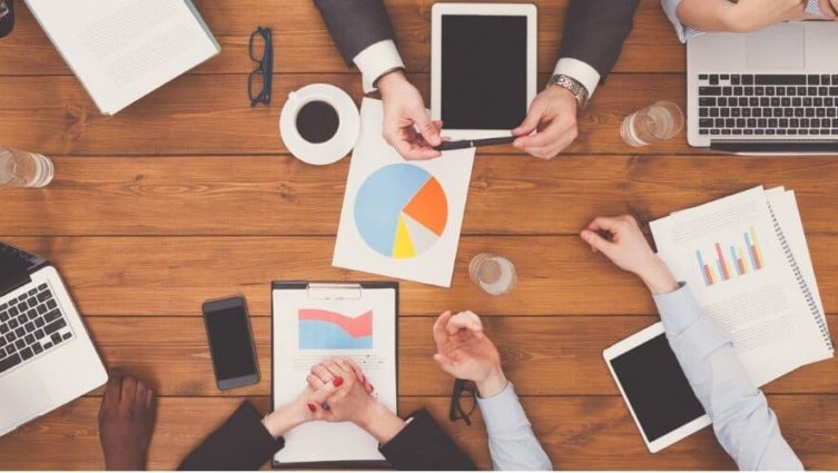 Imagem de uma reunião em ambiente corporativo com as pessoas utilizando laptops, tablets, celulares e gráficos impressos em papel