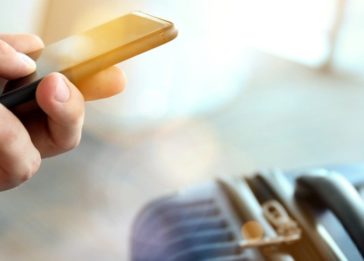 Pessoa no aeroporto mexendo no smartphone e uma mala ao lado