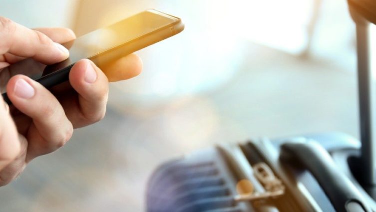 Pessoa no aeroporto mexendo no smartphone e uma mala ao lado