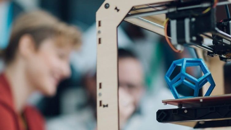 Imagem mostra uma impressora 3D em primeiro plano e pessoa sorrindo no segundo plano.