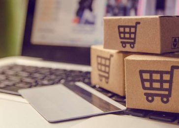 Imagem conceito de um e-commerce: um cartão de crédito está apoiado sobre o teclado de um laptop e, próximo a ele, existem três caixinhas com o ícone de um carrinho de supermercado sobre a superfície