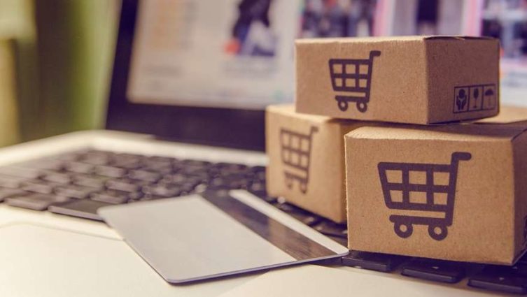 Imagem conceito de um e-commerce: um cartão de crédito está apoiado sobre o teclado de um laptop e, próximo a ele, existem três caixinhas com o ícone de um carrinho de supermercado sobre a superfície