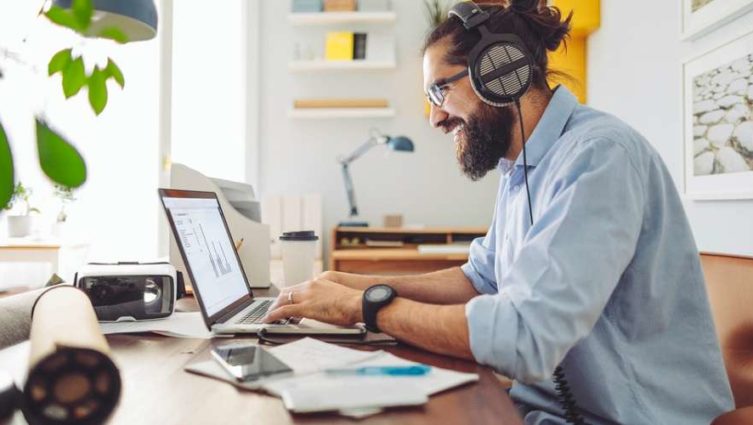 Homem realiza trabalho em casa, a partir de seu laptop, enquanto utiliza um fone de ouvido